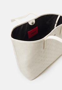 Cremefarvet tote bag med præget logo-mønster, lædermateriale og sort indvendig med et rødt mærke.