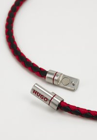 Schwarzes und rotes geflochtenes Lederarmband mit silbernem Metallverschluss. Der Verschluss hat das eingravierte Logo "HUGO" und einen runden Entriegelungsknopf.
