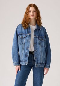 Svetlo modra denim jakna z ohlapnim krojem, s dvema prsna žepa, kovinskimi gumbi in detajli z dvojno šivanko. Kombinirana z belo srajco.