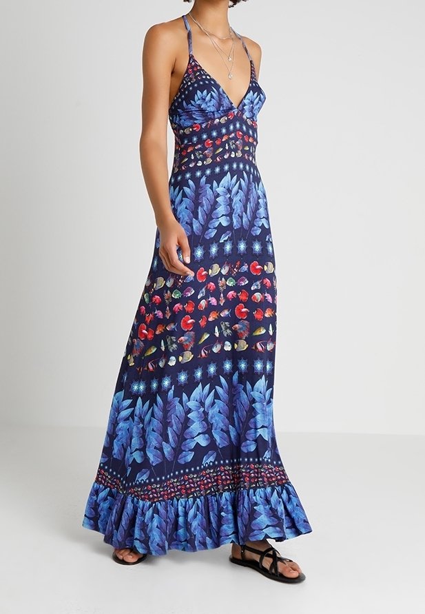 Femme portant une robe maxi sans manches bleu marine avec des motifs de feuilles bleues et de fruits rouges, associée à des sandales plates noires et des colliers superposés.