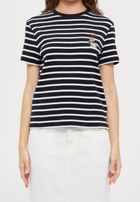 Námořnické a bílé pruhované tričko s krátkým rukávem, kulatým výstřihem a vyšitým designem kočky s červenou mašlí na levé straně hrudi.