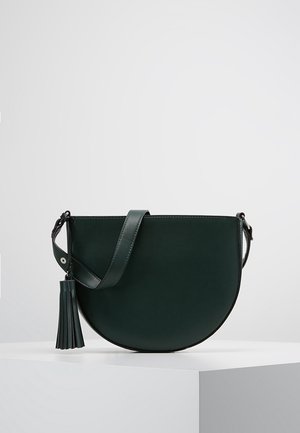 Ciemnozielona, półokrągła torba na ramię z regulowanym paskiem i ozdobą z frędzelków, prezentowana na białym piedestale na jednolitym tle.