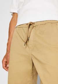 Gros plan sur une main reposant à côté d'un pantalon avec cordon de serrage beige et d'une chemise à manches courtes blanche.