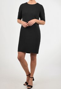 Černé šaty bez rukávů s kulatým výstřihem, hladkým povrchem, po kolena a minimalistickým designem. Nosené s černými sandály na podpatku.