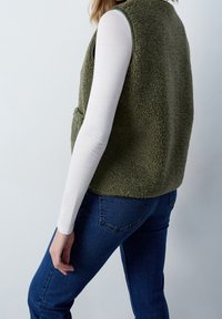 Person, der eine olivgrüne, flauschige Weste über einem weißen Langarmshirt und blauen Jeans tr�ägt, steht vor einem hellgrauen Hintergrund.