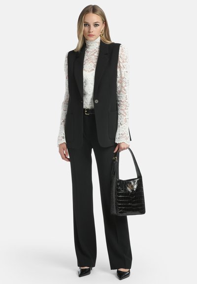 Sort ærmeløs blazer over en hvid blonde top, parret med sorte bukser. Sort patentlæder håndtaske og spidse højhælede sko.