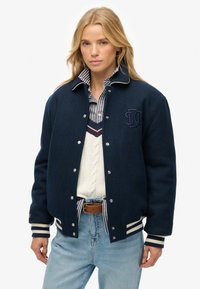 Chaqueta bomber de lana navy con detalles blancos, botones a presión y un logo bordado. Llevada sobre un suéter blanco de punto de cable y una camisa a rayas.