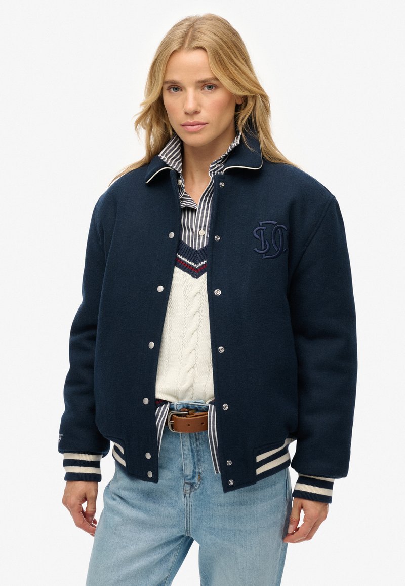 Chaqueta bomber de lana navy con detalles blancos, botones a presión y un logo bordado. Llevada sobre un suéter blanco de punto de cable y una camisa a rayas.