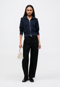 Námořnický pletený svetr na zip, žebrovaná textura, s červenými akcenty na límci; v kombinaci s širokými černými kalhotami a krémovou kabelkou. Černé boty.