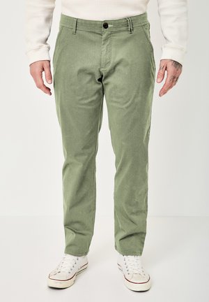 Mann trägt olivgrüne Hosen mit geradem Schnitt und weiße Turnschuhe, steht vor einem schlichten hellen Hintergrund.