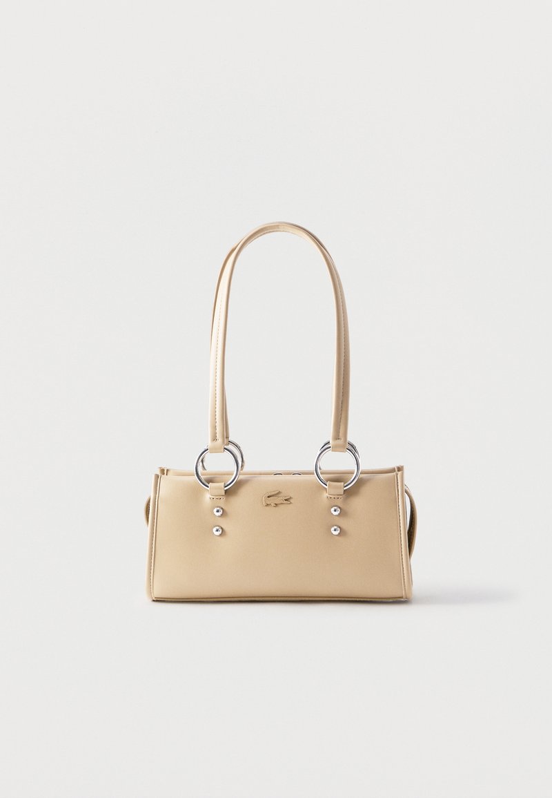 Μπεζ ορθογώνια τσάντα χειρός με διπλές λαβές προσαρτημένες με ασημένιους κρίκους και μικρά ασημένια καρφιά σε λευκό φόντο.