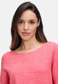 Junge Frau mit langen, dunklen Haaren trägt einen sanften rosa Pullover, schaut frontal mit einem neutralen Gesichtsausdruck vor einem schlichten Hintergrund.