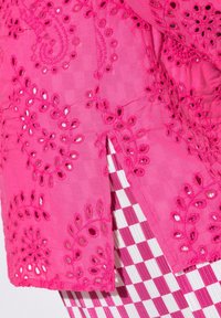 Tessuto fucsia ricamato con motivi floreali e ritagli. Il capo è sovrapposto a un tessuto a quadretti rosa e bianco.
