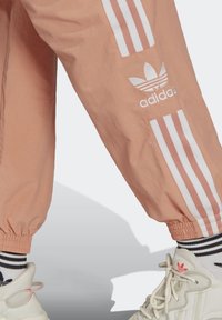 Růžové sportovní kalhoty Adidas s bílými pruhy a logem, s elastickými manžetami a lehkou, zvrásněnou texturou.