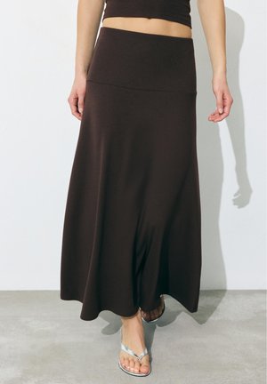 Kobieta w ciemnobrązowej, wysokiej spódnicy do kostek i srebrnych sandałach z odkrytymi palcami, stojąca przy gładkiej białej ścianie.