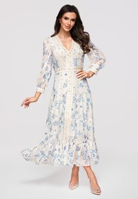 Femme portant une longue robe blanche à fleurs bleues, manches longues transparentes, détails en dentelle et sandales à talons transparents, posant avec la main sur la hanche.