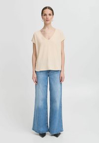 Žena stoji noseći bež majicu s V-izrezom i kratkim rukavima, svijetloplave široke traperice, crne šiljate cipele i naušnice u obliku obruča na bijeloj pozadini.