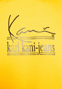 Camisa de algodón amarilla con un logo negro y plateado que exhibe el texto "karl kani-jeans". La tela tiene una apariencia texturizada y acanalada.