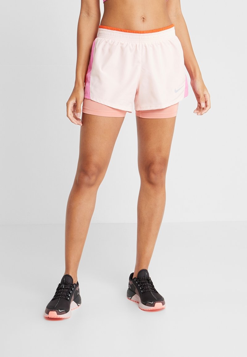 Femme portant un short de course superposé blanc et rose avec une ceinture orange et des chaussures de sport noires avec des semelles roses.