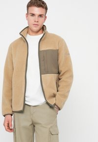 Junger Mann in einer sandfarbenen Fleecejacke mit einer grünen Brusttasche, weißem T-Shirt und beigen Cargo-Hosen, der vor einem schlichten weißen Hintergrund steht.