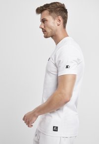 White cotton t-shirt with a crew neckline, short sleeves, and a small black logo on the chest and sleeve. Features a square label on the side.