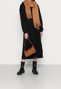 Jupe plissée noire et sweat à capuche court, écharpe frangée marron, sac à main marron brillant et bottines noires à semelles épaisses.