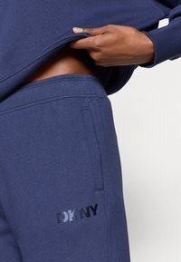 Granatowe spodnie dresowe DKNY o gładkiej teksturze, z boczną kieszenią i czarnym nadrukiem logo na lewym udzie.