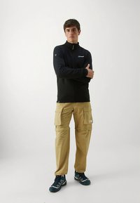 Mustast lukuga fliisist jakk kõrge kraega, koos beežide mitme taskuga cargo-pükstega. Kandmiseks valitud sinised spordijalatsid.