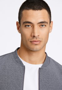 Grå bomberjacka med dragkedja framtill och rundad krage, bärs över en vit t-shirt; slät tyg och rena linjer.