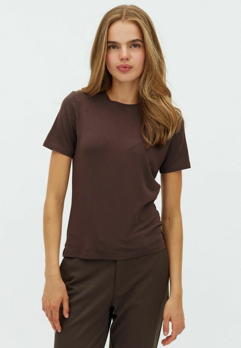Jeune femme aux longs cheveux châtain clair portant un t-shirt uni à manches courtes marron foncé et un pantalon foncé sur un fond clair uni.
