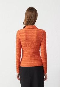 Top arancione a maniche lunghe con texture, colletto e materiale arricciato, dal taglio aderente. Sono parzialmente visibili pantaloni neri.