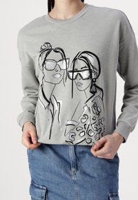 Sudadera gris con un gráfico de dos mujeres con gafas, contorneadas en negro, con un acento de estampado de leopardo. Mangas largas y puños acanalados.