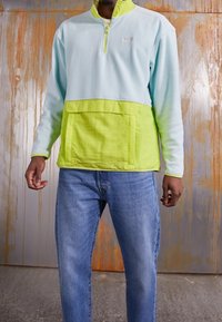 Pullover in pileazzurro con colletto alto e tasca frontale, caratterizzato da un pannello inferiore verde lime e dettagli abbinati. Indossato con jeans blu.