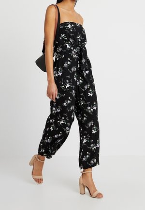 Jumpsuit noir à motifs floraux avec un design sans bretelles, mettant en avant un détail de taille nouée, des motifs floraux et un pantalon décontracté. Les sandales sont de couleur beige clair.