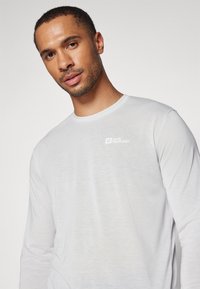 Langärmliges weißes T-Shirt aus leichtem Stoff mit rundem Halsausschnitt und kleinem Logo auf der linken Brust. Glatte Textur, minimalistisches Design.