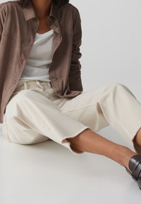 Personne assise portant une veste marron boutonnée, un t-shirt blanc, un jean large aux jambes larges couleur écru avec des ourlets bruts, et des chaussures noires à enfiler sur un fond uni.