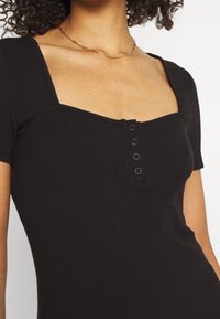 Svart ribbad topp med korta ärmar, fyrkantig halsringning och tre svarta knapppdetaljer framtill. Slät textur och åtsittande passform.