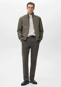 Chaqueta bomber caqui con cuello alto, combinada con un suéter gris claro, pantalones gris ajustados y zapatos de cuero negros.