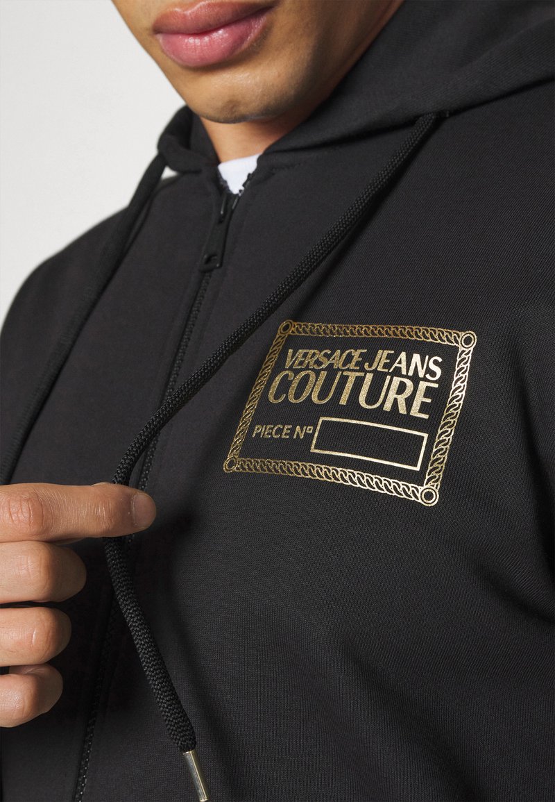 Zwarte zip-up hoodie met gouden logo patch, verstelbare trekkoord en textuur materiaal. Bevat een close-up van het logo en details van de rits.
