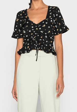 Kobieta w czarnej, kwiecistej bluzce z marszczeniami i krótkimi bufkami, w jasnych, wysokich spodniach, stojąca na jednolitym tle.