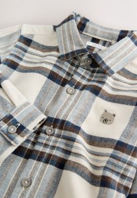 Chemise en flanelle à carreaux bleu clair, crème et marron. Elle présente un col, une patte de boutonnage à l'avant et un logo ours sur la poche poitrine.