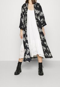 Kimono s černobílým vzorem přes bílé šaty, zdobené krajkovými detaily na lemu, a černé šněrovací boty s masivní podrážkou.