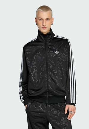 Młody mężczyzna z krótkimi blond włosami, ubrany w czarną dresową kurtkę i spodnie Adidas z subtelnymi wzorami w stylu komiksowym oraz białymi paskami na rękawach.