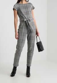 Šedý kostkovaný overal s krátkými rukávy, stahovaným pasem a bočními kapsami, doplněný černými kotníkovými botami a malou černou kabelkou.