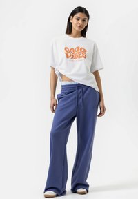 Witte oversized t-shirt met oranje opdruk "GOOD VIBES STATE OF MIND", gecombineerd met blauwe wijde sweatpants en witte sneakers.