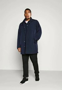 Laivastonsininen pitkä sadetakki, valmistettu sileästä materiaalista, jossa on sivutaskut ja nappikiinnitys, yhdistetty mustiin housuihin ja lenkkareihin.