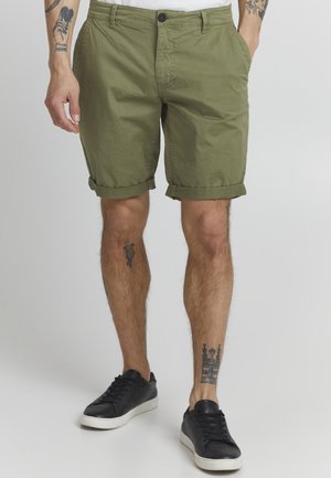 Olivenfarget bomulls-shorts med oppbrettede ermer, med sidelommer og knappefeste, brukt med svarte joggesko.