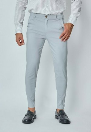 Pantalon ajusté gris clair avec une texture lisse, doté d'une fermeture à bouton et de poches latérales, associé à des mocassins noirs.