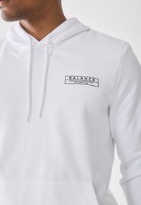 Mand iført hvid hættetrøje med frontlomme og teksten "BALANCE Everything is falling into place as it’s meant to be" på brystet.