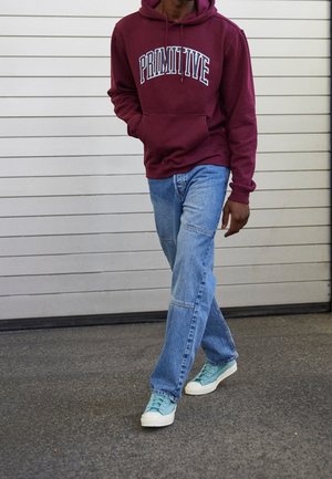 Person, der einen dunkelroten "Primitive" Hoodie, hellblaue Jeans und türkisfarbene Sneakers trägt, vor einem weißen, mit Paneelen versehenen Wandhintergrund.
